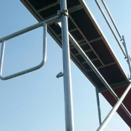 Metalowe rusztowanie z drewnianym pomostem roboczym na tle błękitnego nieba, widok z dołu.