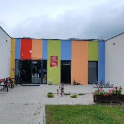 Nowoczesny budynek przedszkola z kolorową fasadą w pionowe pasy: czerwony, pomarańczowy, żółty, zielony i niebieski. Widoczne wejście z przeszklonymi drzwiami, ławki i donice z kwiatami. Tabliczka...