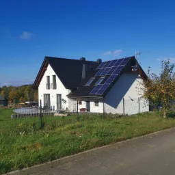 Biały dom jednorodzinny z czarnym dachem i panelami słonecznymi na południowej połaci, widziany z poziomu ulicy za ogrodzeniem z siatki.