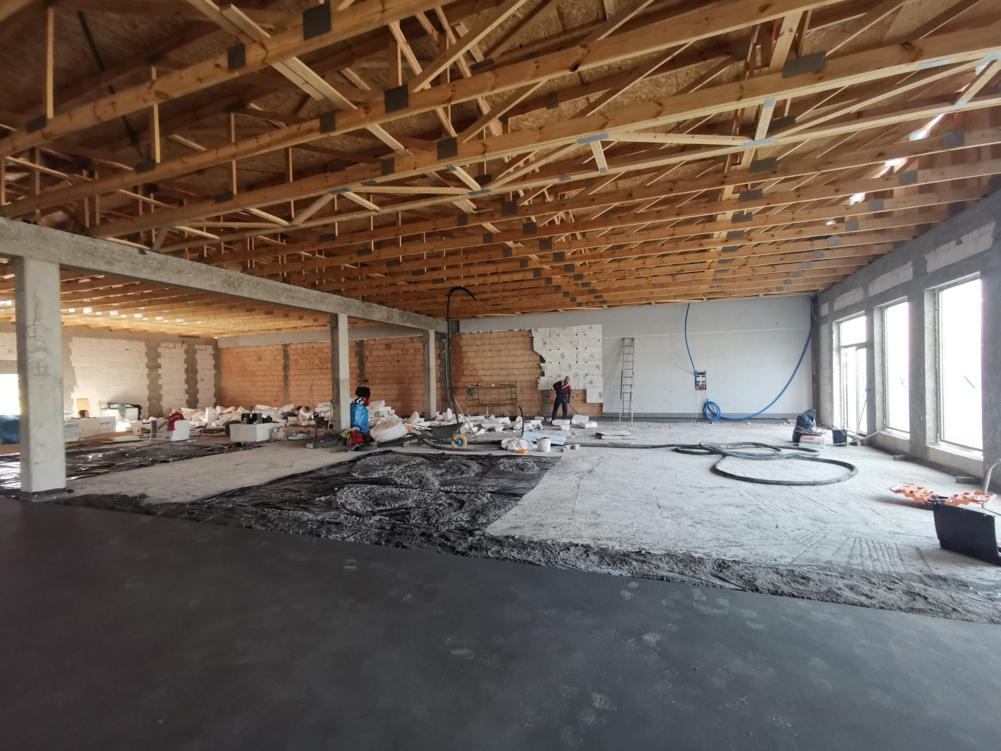 Wnętrze budynku w trakcie prac budowlanych: widoczne drewniane belki stropowe, murowane ściany, wylewka podłogowa w trakcie wykonywania, materiały budowlane oraz pracownicy w oddali.