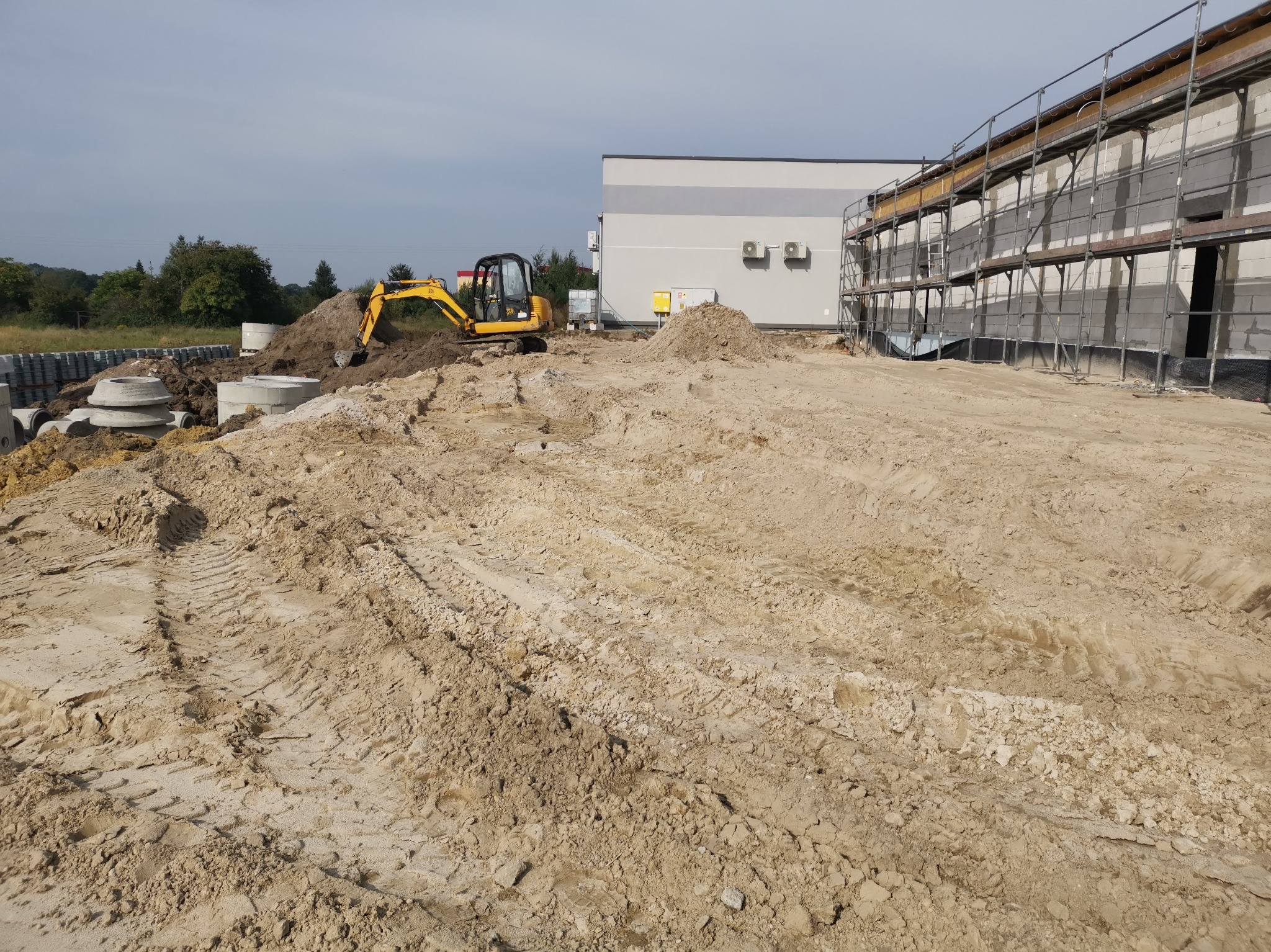 Żółta minikoparka na placu budowy z przygotowanym terenem pod fundamenty, obok składowane betonowe kręgi, w tle budynek w trakcie wznoszenia z rusztowaniem.