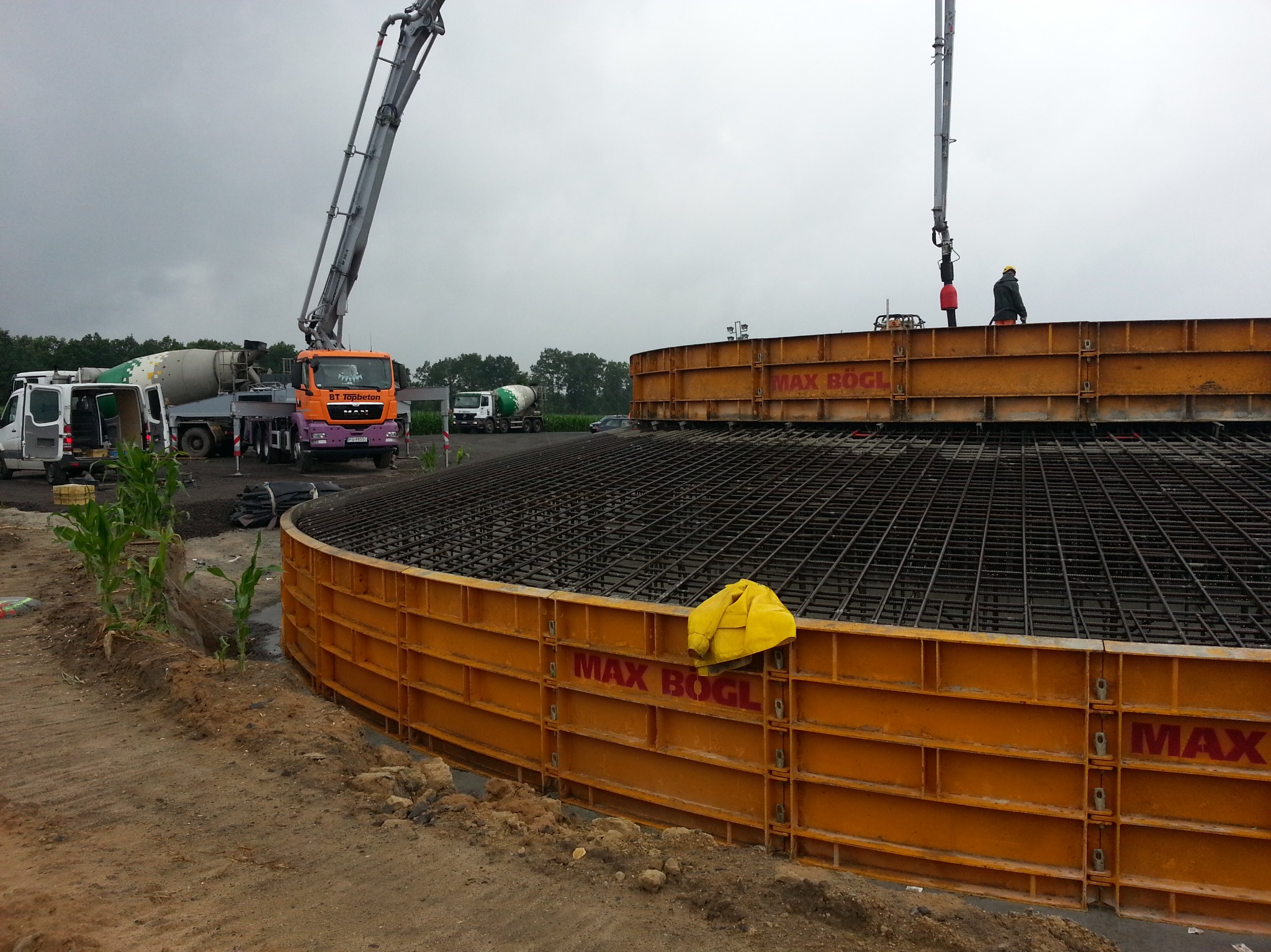 Budowa okrągłego zbiornika z użyciem szalunków systemowych Max Bögl, widoczna zbrojona siatka stalowa, pompy do betonu i betonomieszarki w tle, pochmurne niebo.