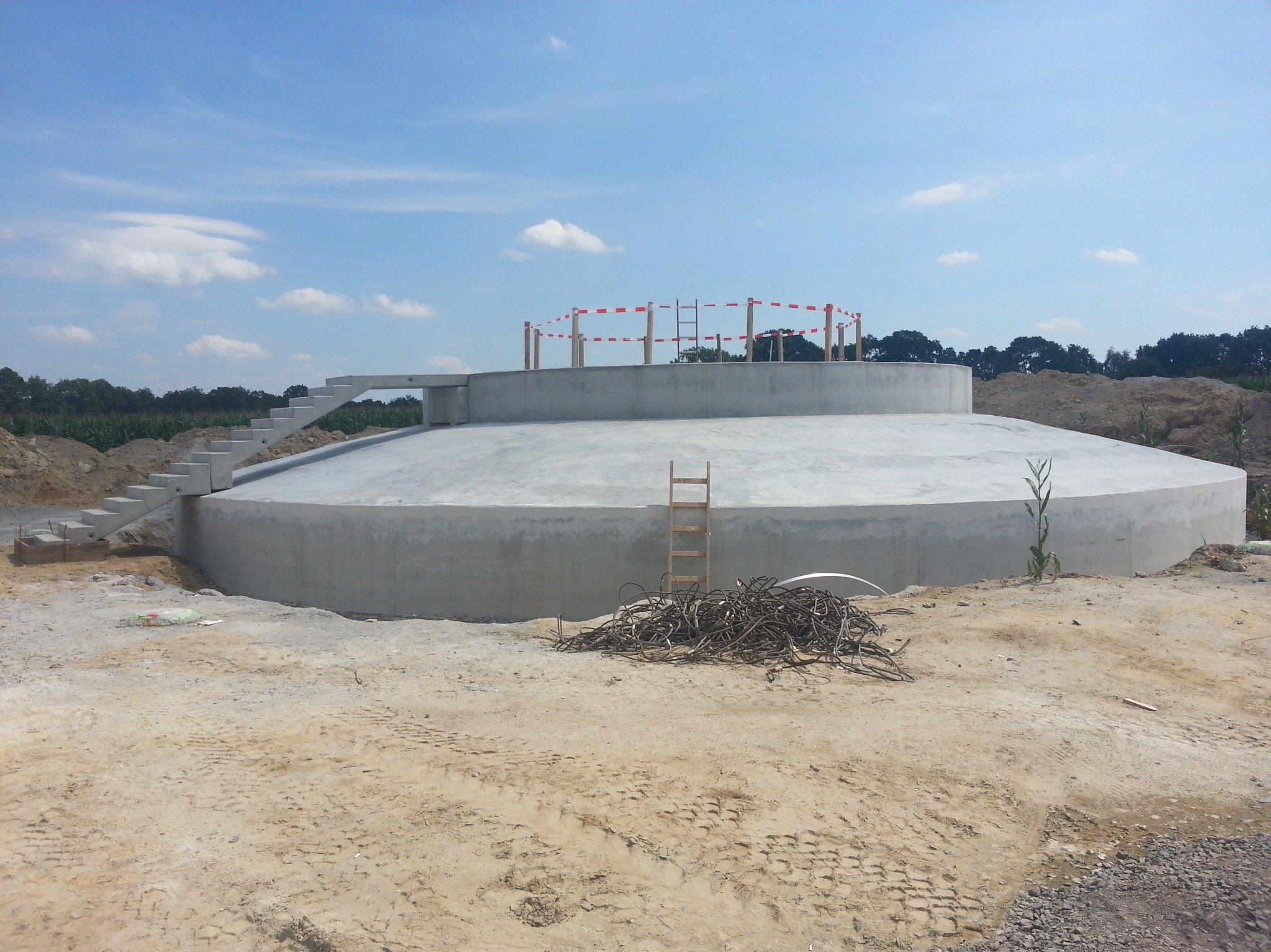 Betonowy zbiornik retencyjny w trakcie budowy, widoczne schody, drabina na górze zbiornika i sterta zbrojenia na placu budowy