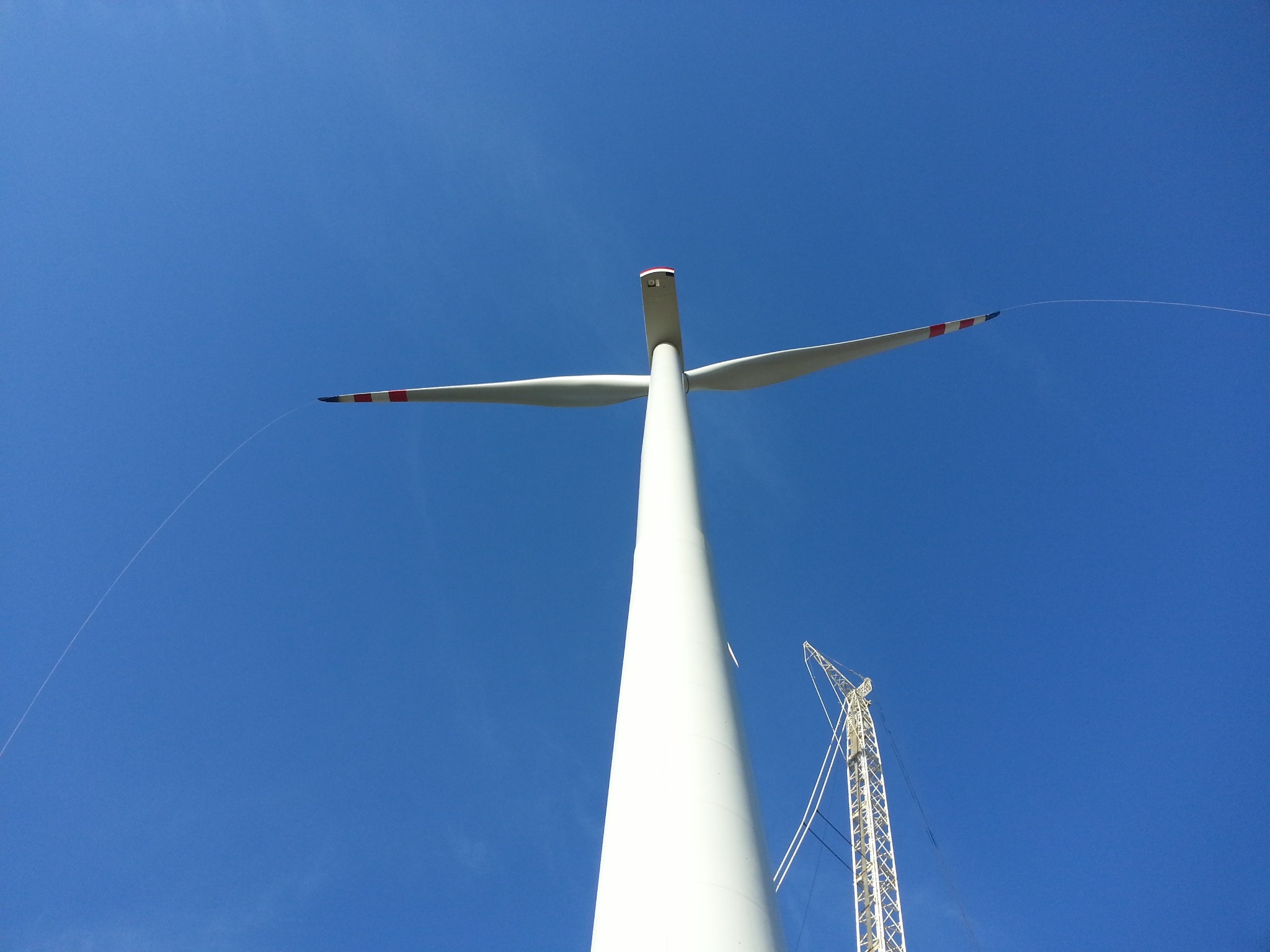 Wysoka biała turbina wiatrowa widziana z dołu na tle błękitnego nieba, z widocznym fragmentem dźwigu w prawym dolnym rogu.