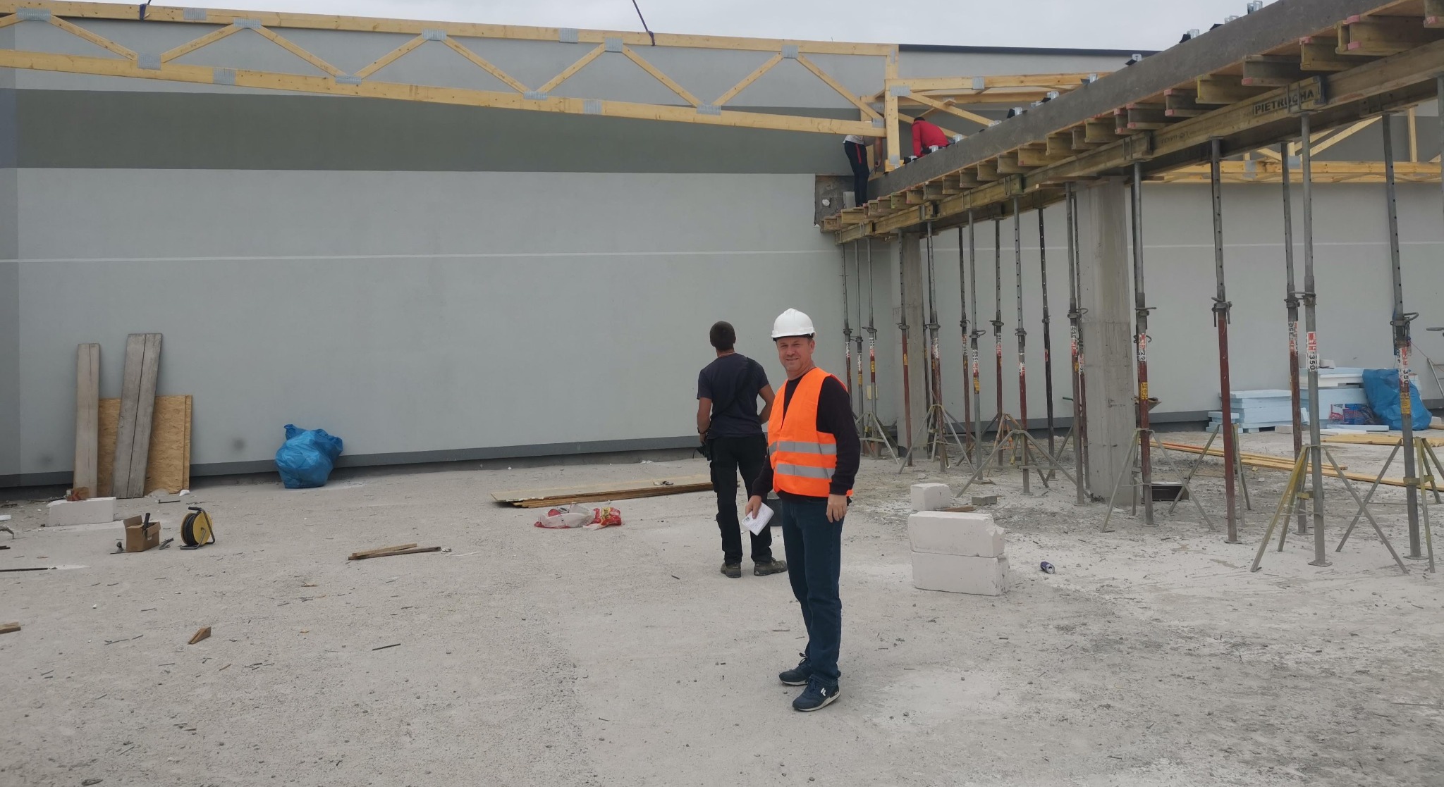 Budowa hali: pracownik w kamizelce odblaskowej i kasku na tle betonowych słupów podtrzymujących szalunek stropu. W tle widoczna szara ściana i drewniana konstrukcja dachu.