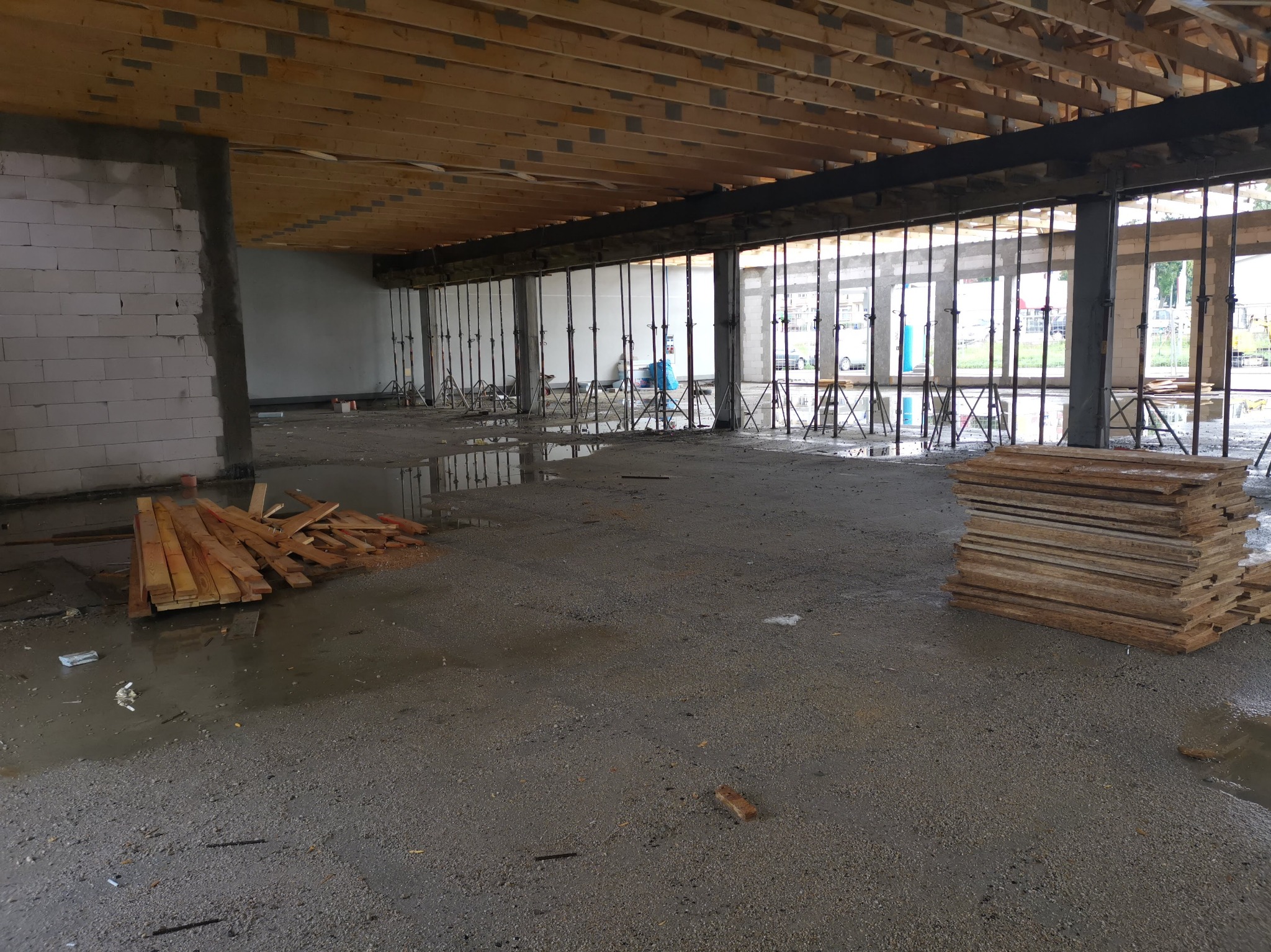 Wnętrze surowego budynku w trakcie budowy: widoczne drewniane belki stropowe podparte metalowymi stemplami, mokra betonowa podłoga z kałużami i stosami desek.