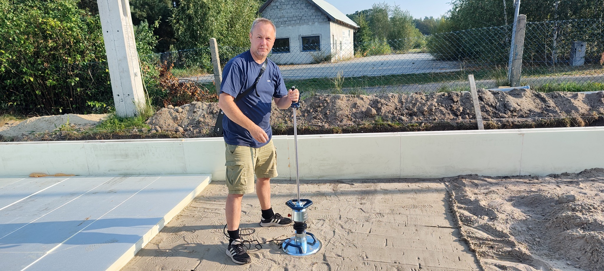 Mężczyzna obsługuje zagęszczarkę na piaszczystym podłożu wewnątrz fundamentów budynku, widoczne białe płyty izolacyjne i przygotowany teren pod wylewkę.