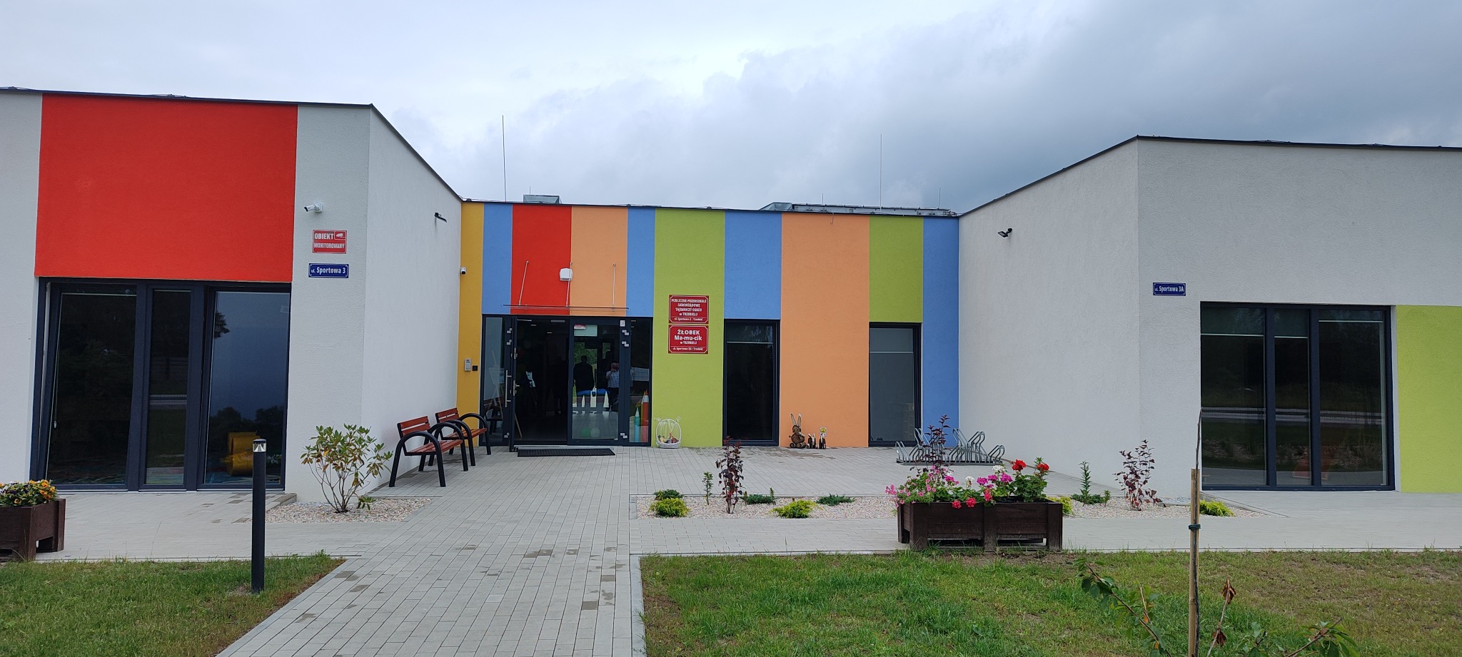 Nowoczesny budynek przedszkola z kolorową fasadą w pionowe pasy: czerwony, pomarańczowy, żółty, zielony i niebieski. Widoczne wejście z przeszklonymi drzwiami, ławki i donice z kwiatami. Tabliczka...