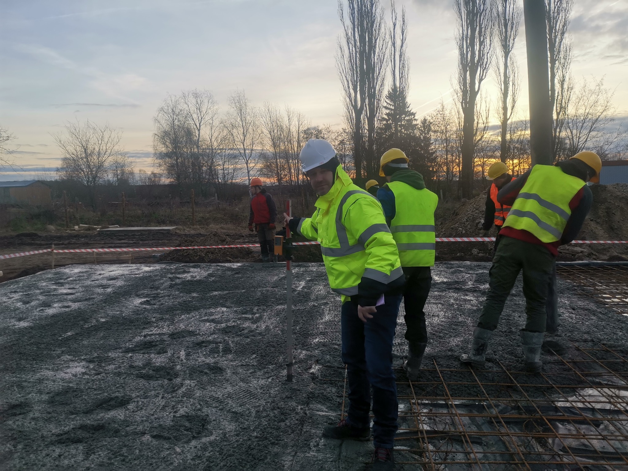 Pracownicy w odblaskowych kamizelkach i kaskach pracują na budowie, poziomując wylewkę pod fundamenty na siatce zbrojeniowej, przy zachodzącym słońcu.