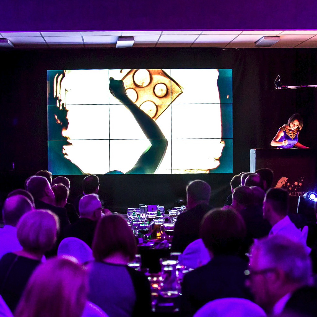 Iluzjonistka prezentuje pokaz rysowania piaskiem na dużym ekranie podczas imprezy firmowej, publiczność siedzi przy stołach.