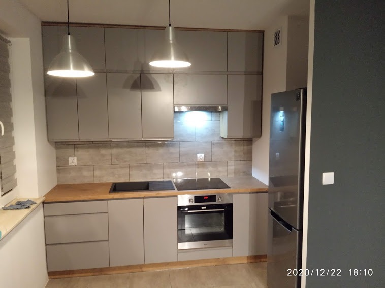 Szara zabudowa kuchenna z drewnianym blatem, srebrna lodówka i dwa wiszące metalowe klosze lamp, widok na fragment ściany pomalowanej na ciemny kolor.