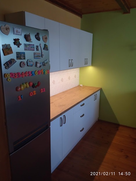 Nowoczesna kuchnia z białymi szafkami i drewnianym blatem, ozdobiona magnesami na lodówce, w tle zielona ściana.