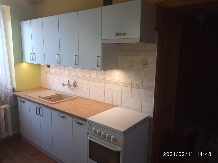 Jasna kuchnia z białymi szafkami, drewnianym blatem i starą, białą kuchenką; widok na kafelki i baterię umywalkową.