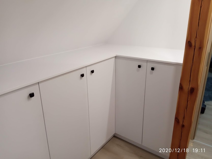 Białe szafki z czarnymi uchwytami, zabudowa pod skosem na poddaszu, widoczna drewniana belka konstrukcyjna.