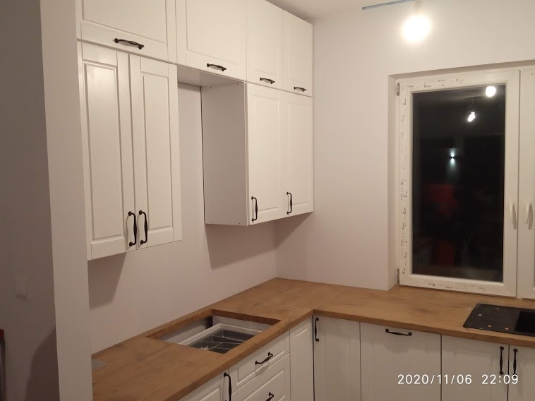 Fragment białej kuchni z drewnianym blatem, widoczny zlew z ociekaczem i płyta indukcyjna przy oknie.