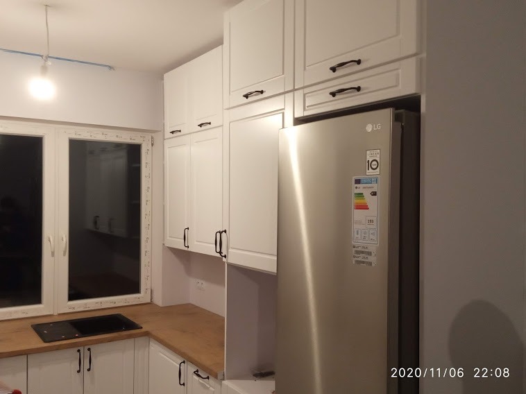 Biała zabudowa kuchenna z drewnianym blatem, czarnymi uchwytami i srebrną lodówką LG w tle okna.