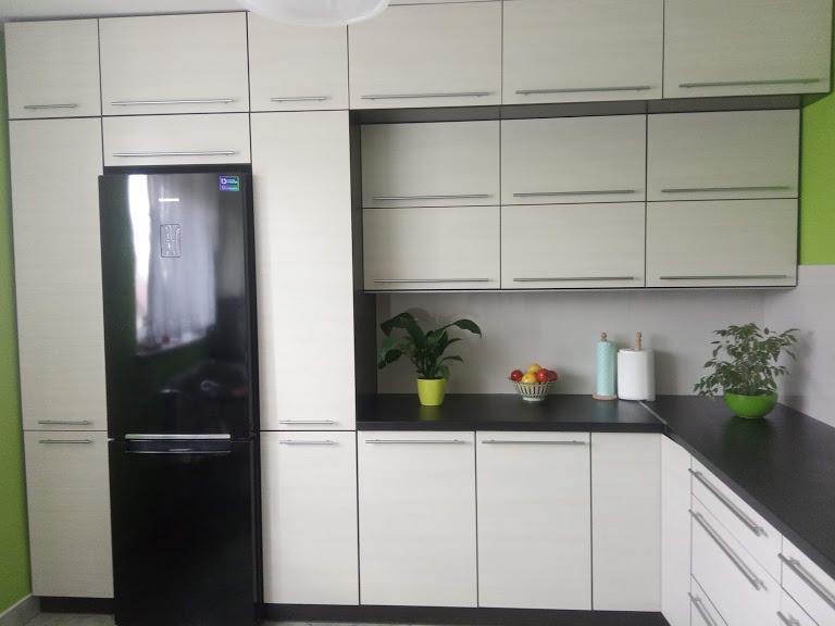 Jasna zabudowa kuchenna z frontami w odcieniach bieli i beżu, z czarnym blatem i czarną lodówką Samsung wkomponowaną w szafki, uzupełniona zielonymi roślinami doniczkowymi i misą z owocami.