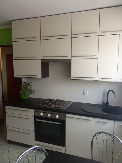 Jasna zabudowa kuchenna z górnymi szafkami z poziomymi uchwytami, czarnym blatem i sprzętem AGD, widok na fragment stołu i krzesła.