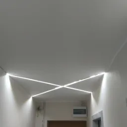 Korytarz z nowoczesnym oświetleniem LED na suficie, tworzącym geometryczny wzór krzyżujących się linii świetlnych. Widoczne przewody elektryczne na ścianie obok drewnianych drzwi wejściowych.