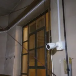 Kamera monitoringu zamontowana na ścianie obok okna z ciemnymi ramami, widoczne przewody i puszki instalacyjne.