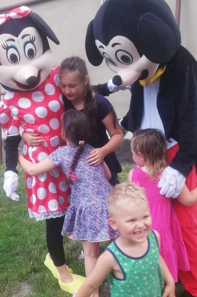 Dzieci przytulają się do postaci Myszki Miki i Minnie w kostiumach na tle zielonej trawy, mały chłopiec z przodu uśmiecha się szeroko, postać Minnie ma na nogach żółte, płaskie buty.