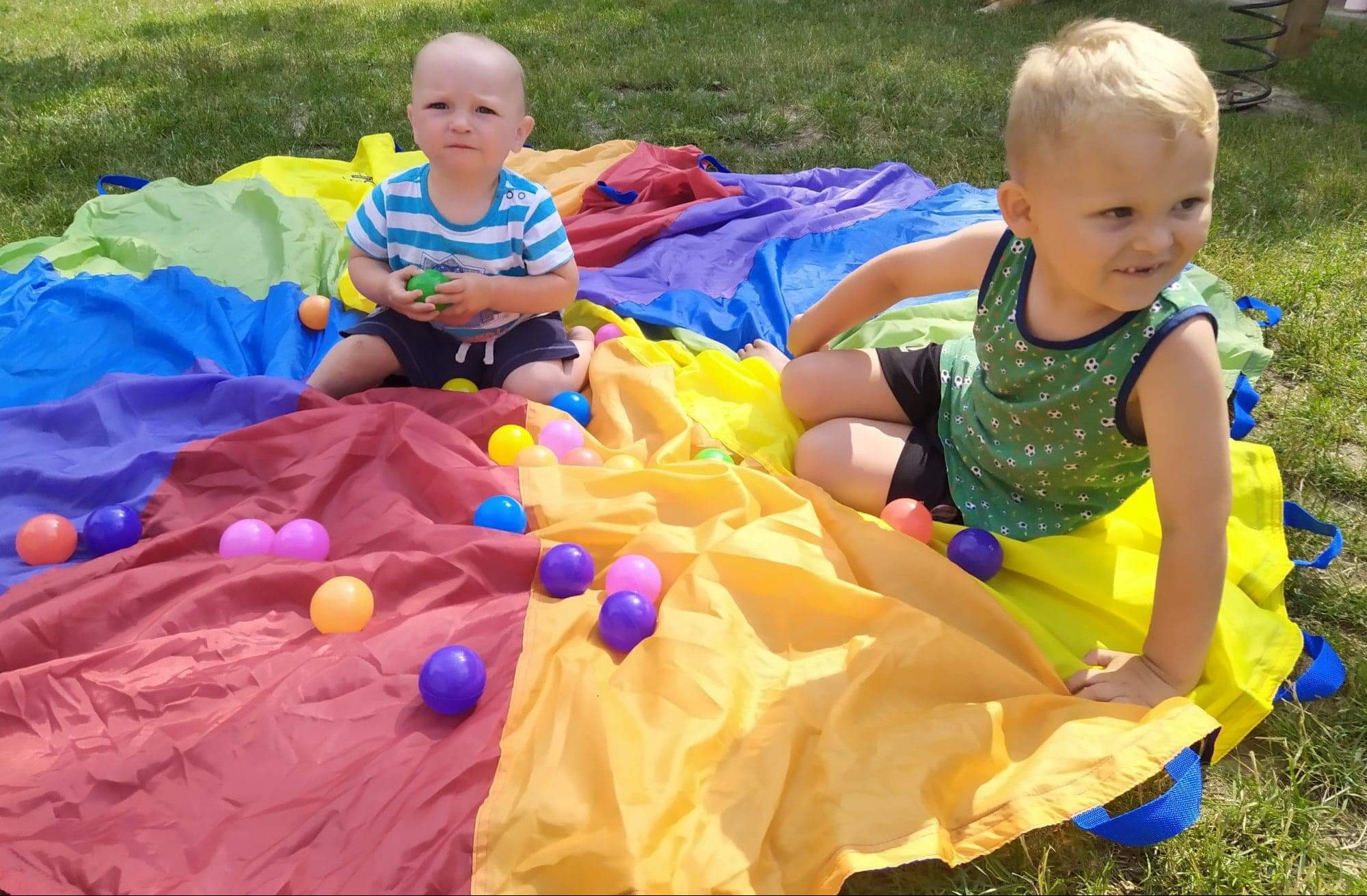 Dwaj chłopcy bawią się na kolorowej chuście animacyjnej z rozrzuconymi piłeczkami na trawie.