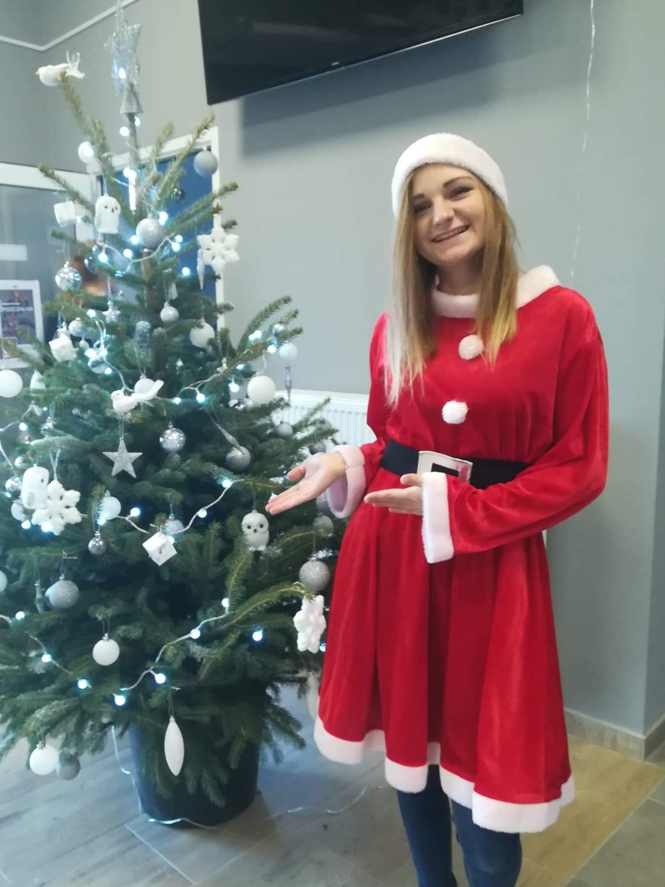 Kobieta w stroju Mikołajki, z uśmiechem, prezentuje udekorowaną choinkę z białymi ozdobami i światełkami w pomieszczeniu z szarą ścianą.