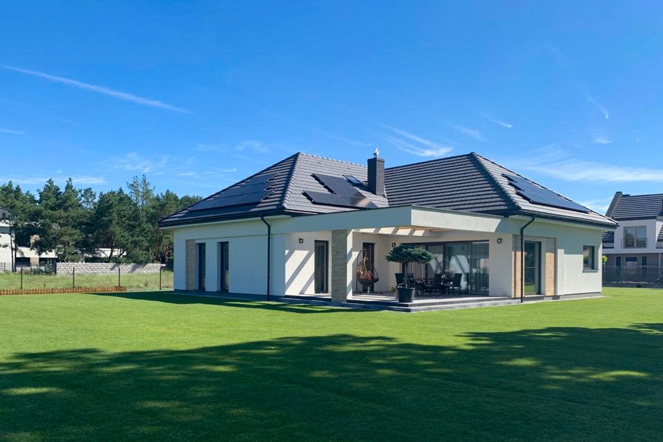 Nowoczesny dom jednorodzinny z ciemną dachówką i panelami słonecznymi na dachu, duży trawnik, błękitne niebo