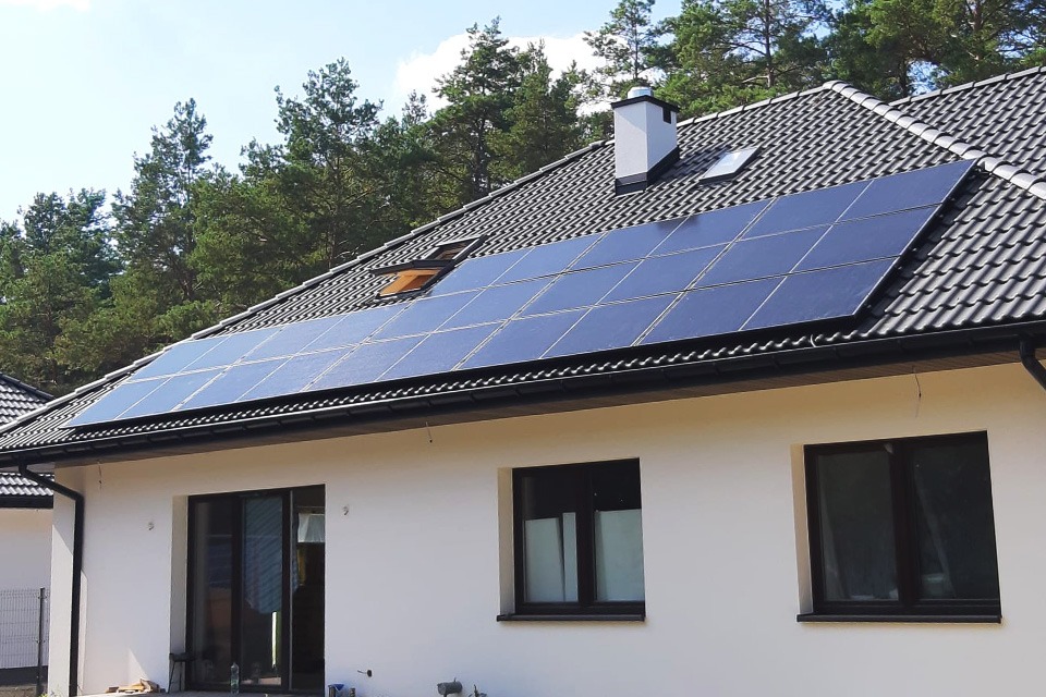 Panele fotowoltaiczne na dachu domu jednorodzinnego z czarną dachówką, widoczne okno dachowe i komin, tło stanowią drzewa.