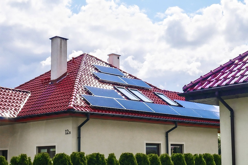 Czerwony dach z panelami słonecznymi na jasnym budynku z numerem 12b, widok z dołu na tle błękitnego nieba z chmurami.
