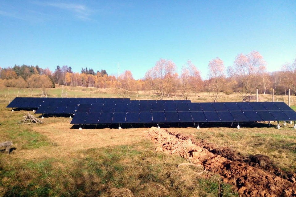 Rozległa instalacja paneli słonecznych na łące, widoczny wykop w ziemi, w tle drzewa z jesiennymi liśćmi i błękitne niebo.
