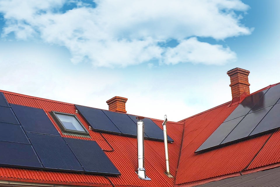 Czerwony dach z panelami słonecznymi, kominami z cegły i oknem dachowym na tle błękitnego nieba z chmurami.
