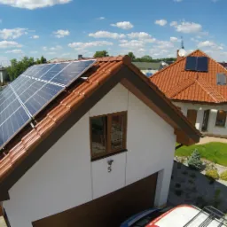 Dom jednorodzinny z panelami fotowoltaicznymi na dachu z dachówki ceramicznej, widok z góry na instalację i otoczenie posesji w słoneczny dzień.