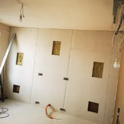 Montaż płyt kartonowo-gipsowych na ścianie z widocznymi otworami na instalacje, przewody elektryczne zabezpieczone peszlem, surowy stan wykończenia, drabina oparta o ścianę.