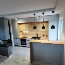 Nowoczesna kuchnia z wyspą i kominkiem, szaro-białe szafki z drewnianymi akcentami, marmurowe podłogi, wiszące czarne lampy.