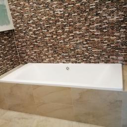 Ideal-bud Paweł Okhovych Firma remontowo-wykończeniowa - Biała wanna obudowana beżowymi płytkami, ściana wyłożona mozaiką z brązowych i beżowych elementów, widoczny grzejnik drabinkowy i fragment ściany w trakcie wykończenia.