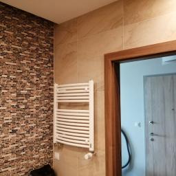 Ideal-bud Paweł Okhovych Firma remontowo-wykończeniowa - Wykończona ściana łazienki z mozaiką i grzejnikiem drabinkowym, widok na otwarte drzwi do innego pomieszczenia.