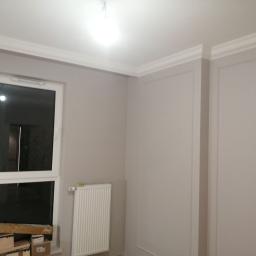 Ideal-bud Paweł Okhovych Firma remontowo-wykończeniowa - Pokój po malowaniu z białymi listwami dekoracyjnymi na ścianach, białym grzejnikiem i oknem, w którym leżą kartony.