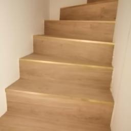 Ideal-bud Paweł Okhovych Firma remontowo-wykończeniowa - Schody wykończone panelami z drewnopodobnym wzorem i złotymi listwami krawędziowymi, widok z dołu.