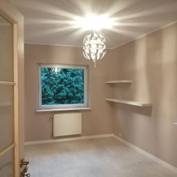 Ideal-bud Paweł Okhovych Firma remontowo-wykończeniowa - Pomalowane na beżowo wnętrze pokoju z jasną podłogą, oknem z widokiem na zieleń, białym grzejnikiem i designerską lampą sufitową.