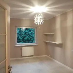 Pomalowane na beżowo wnętrze pokoju z jasną podłogą, oknem z widokiem na zieleń, białym grzejnikiem i designerską lampą sufitową.