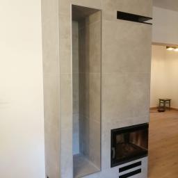 Ideal-bud Paweł Okhovych Firma remontowo-wykończeniowa - Obudowa kominka wykończona płytkami ceramicznymi w odcieniach szarości, z wnęką na drewno i czarnymi elementami dekoracyjnymi, na tle drewnianej podłogi i fragmentu białej ściany.