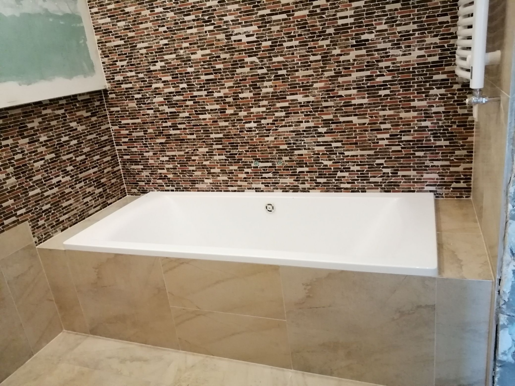 Biała wanna obudowana beżowymi płytkami, ściana wyłożona mozaiką z brązowych i beżowych elementów, widoczny grzejnik drabinkowy i fragment ściany w trakcie wykończenia.
