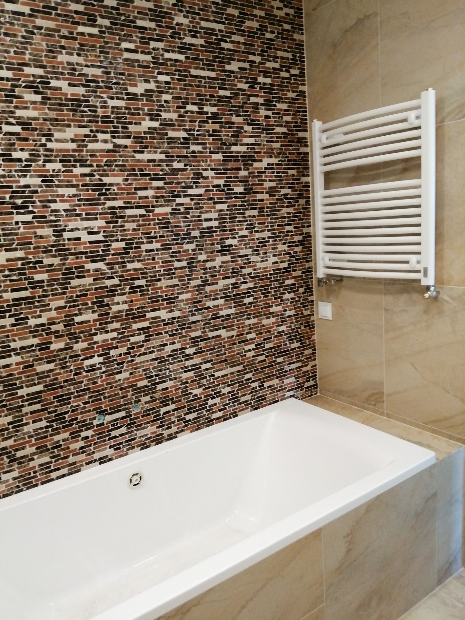 Biała wanna z mozaiką w odcieniach brązu i czerni na ścianie, obok grzejnik drabinkowy i płytki imitujące marmur, widok z góry.