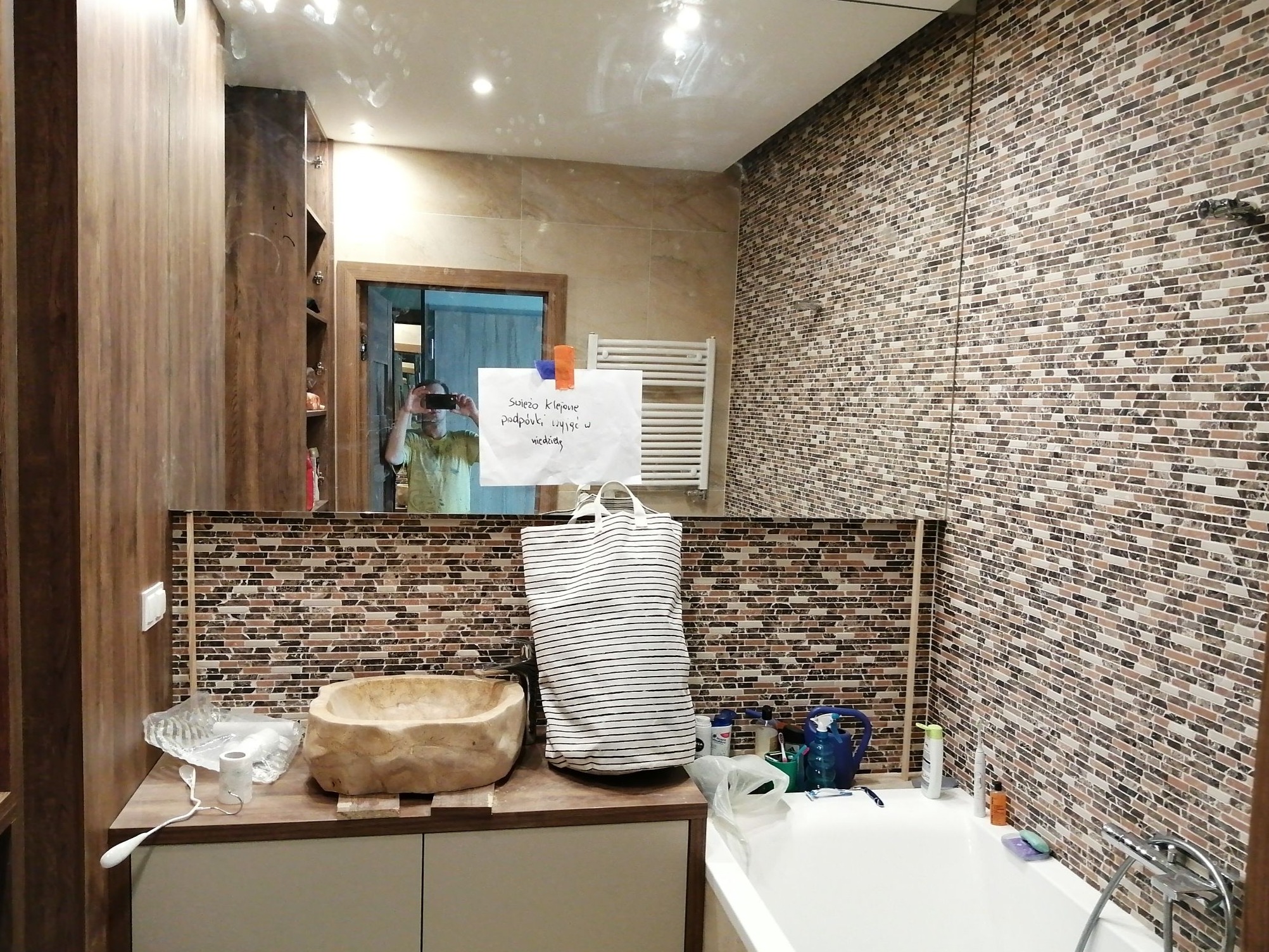 Umywalka z kamienia w łazience z mozaikową ścianą, torba w paski i kartka 'świeżo klejone podpórki, używać w niedzielę' przyklejona do lustra.