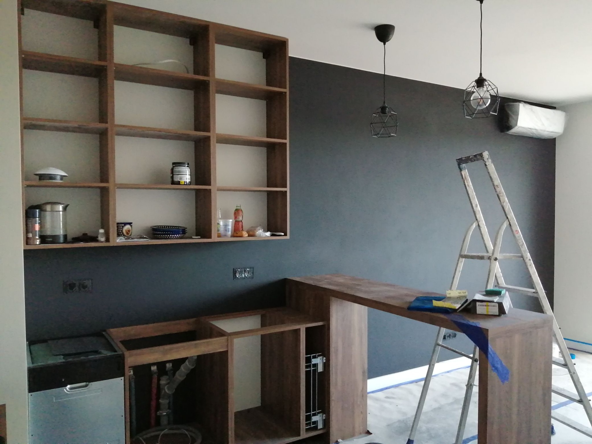 Wnętrze w trakcie wykańczania: drewniane szafki kuchenne bez frontów, półki z wyposażeniem, drabina oparta o ciemną ścianę, wiszące lampy i klimatyzator.