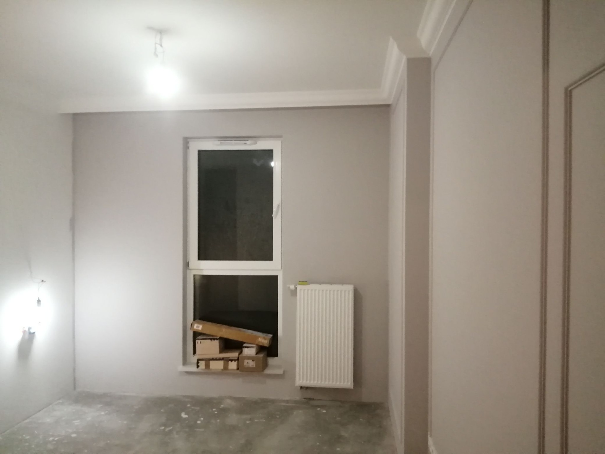 Pokój w trakcie remontu: świeżo pomalowane ściany na szaro, białe listwy przysufitowe, grzejnik i okno z kartonami na parapecie, surowa podłoga, widoczny kabel od lampy