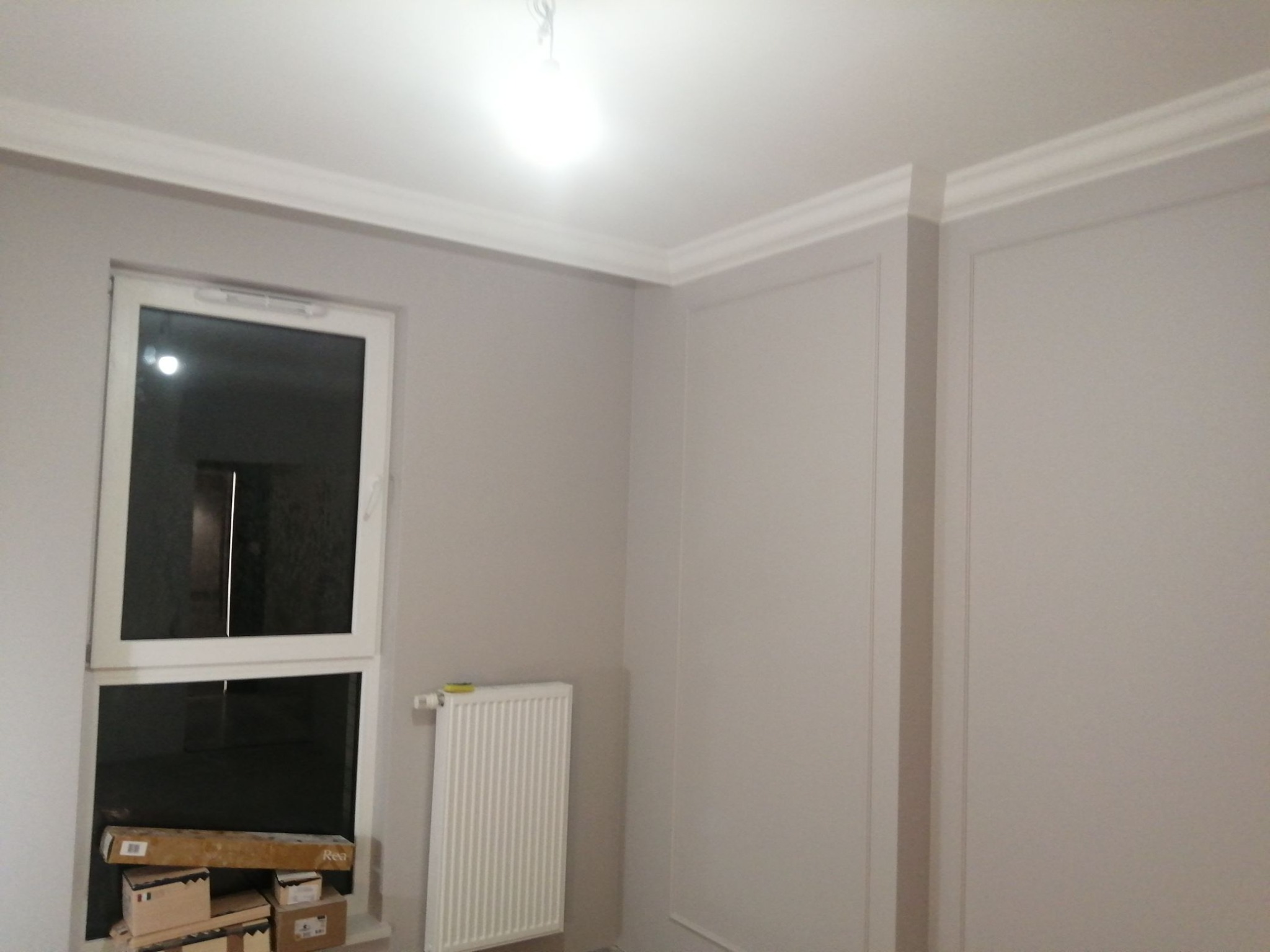 Pokój po malowaniu z białymi listwami dekoracyjnymi na ścianach, białym grzejnikiem i oknem, w którym leżą kartony.
