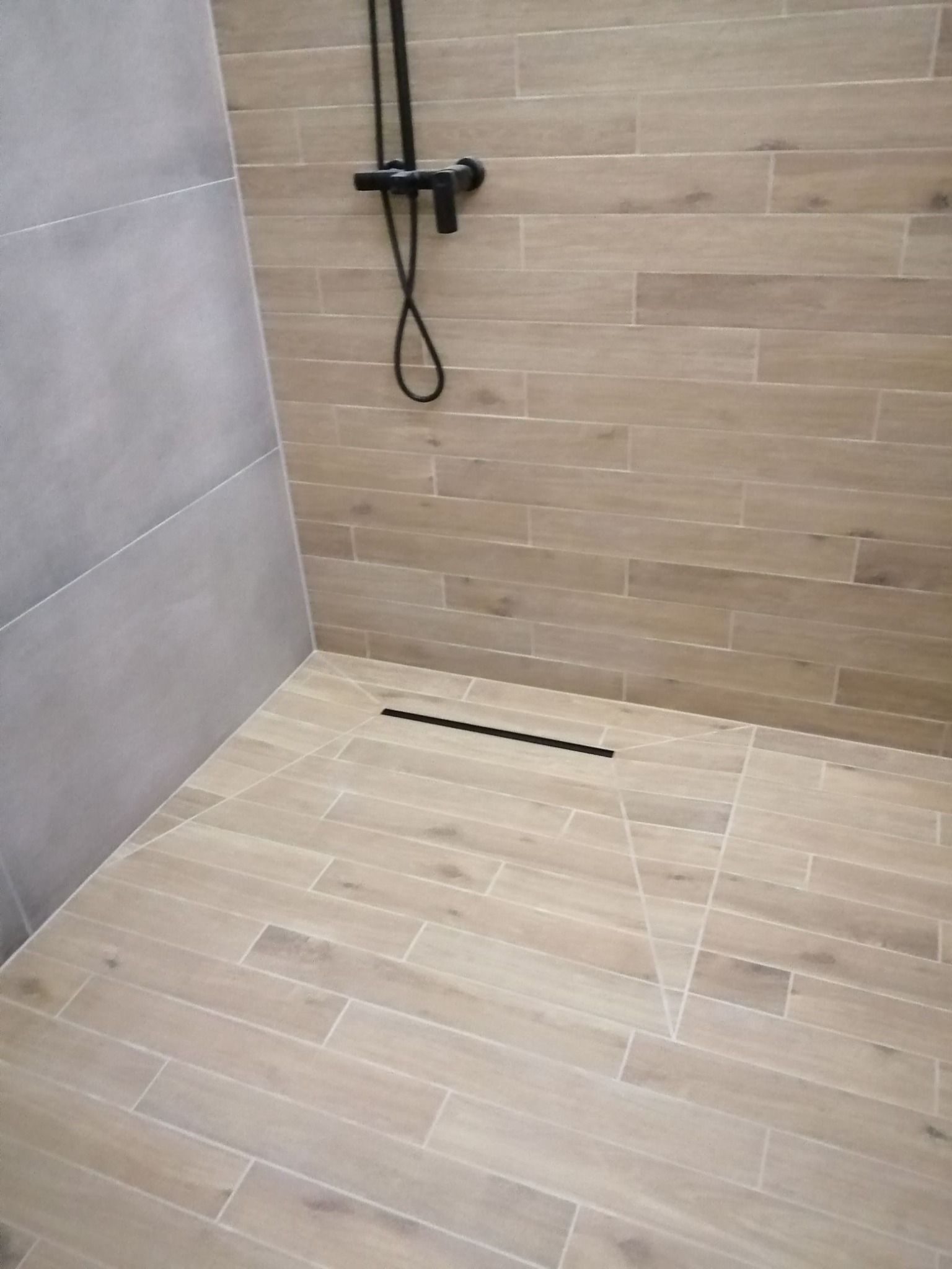 Wykończona kabina prysznicowa z odpływem liniowym i armaturą w kolorze czarnym, wyłożona płytkami imitującymi drewno, z szarą ścianą po lewej stronie.