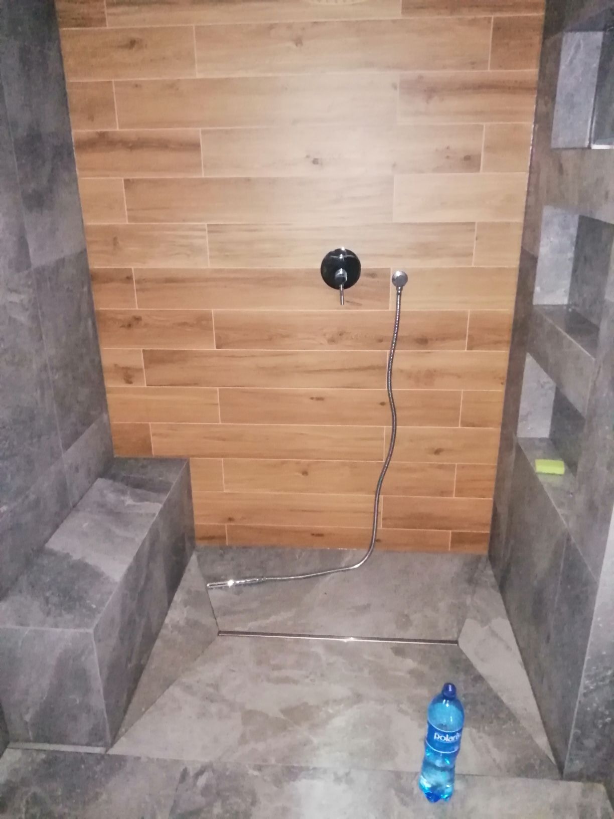 Nowoczesny prysznic walk-in z drewnopodobnymi płytkami na ścianie, szarą strefą siedziska i odpływem liniowym w podłodze; butelka wody na podłodze wskazuje na trwające prace wykończeniowe.